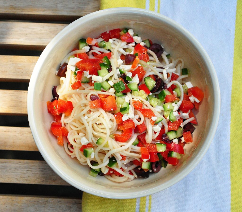 Noodles with Greek Salad ingredients in a bowl on a yellow striped dish towel
