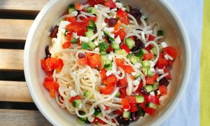 Noodles with Greek Salad ingredients in a bowl on a yellow striped dish towel