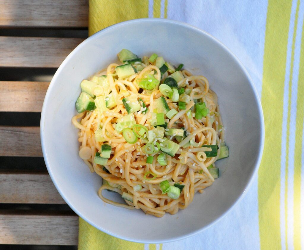 white bowl with sesame noodles topped with cucumbers and scallions