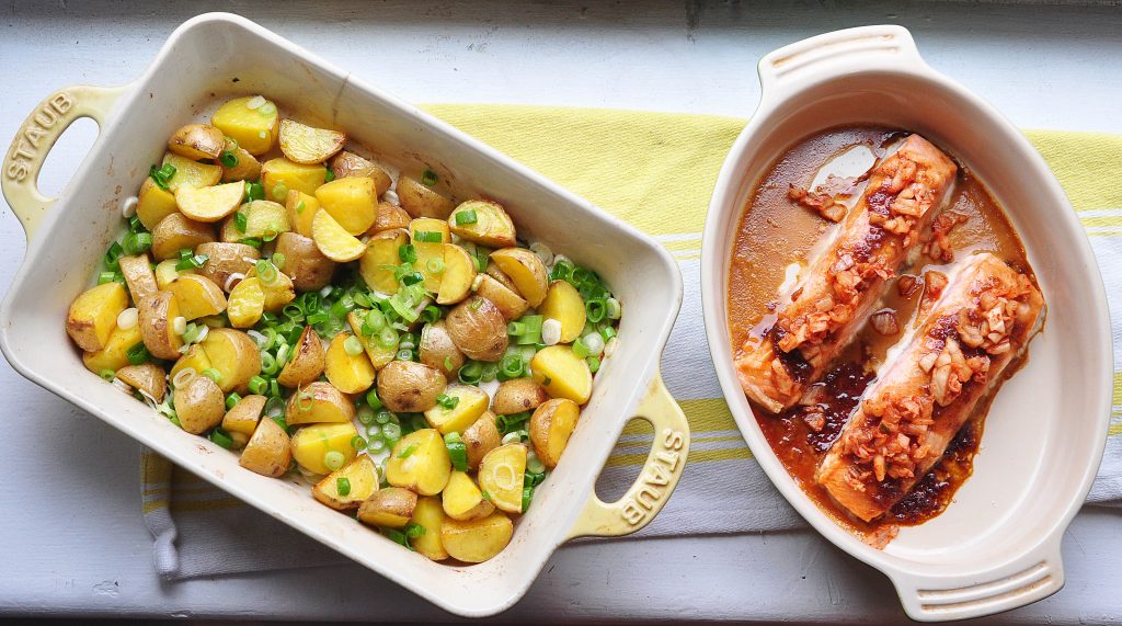 two casserole dishes side by side. One with roasted potatoes and scallions, the other with soy-garlic glazed salmon topped with kimchi.