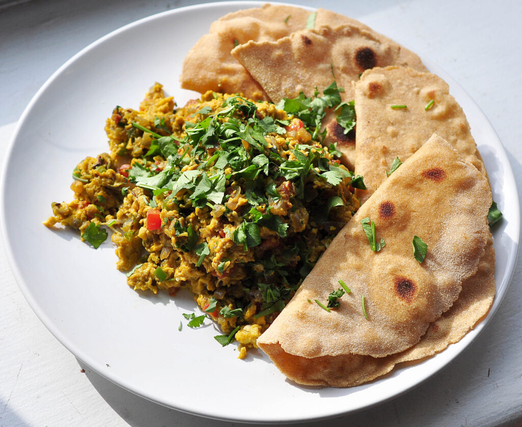 white plate with pile of very yellow scrambled eggs and vegetables with cilantro on top and four pieces of roti