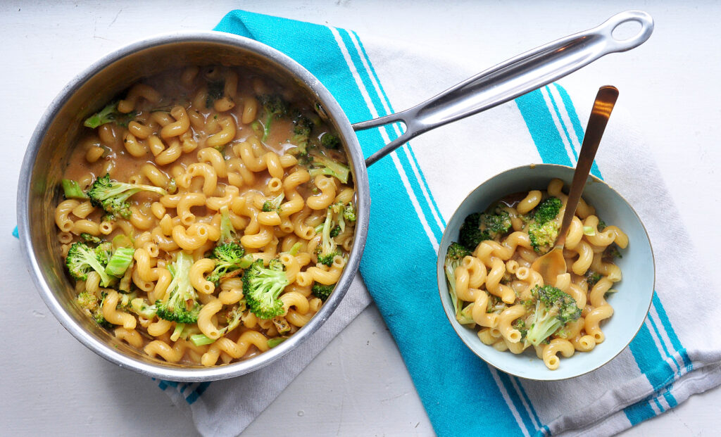 welsh rarebit macaroni and cheese with broccoli in a pot on a teal striped tea towel and in a bowl with a fork