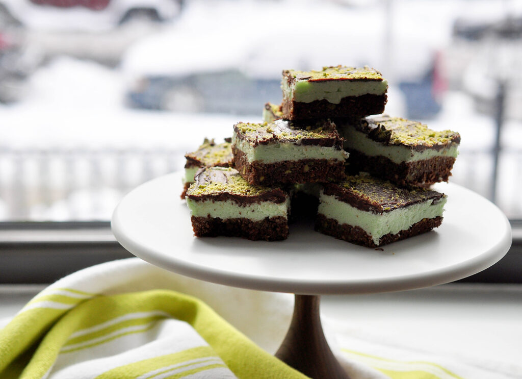 pistachio nanaimo bars piled on a cake platter by a window