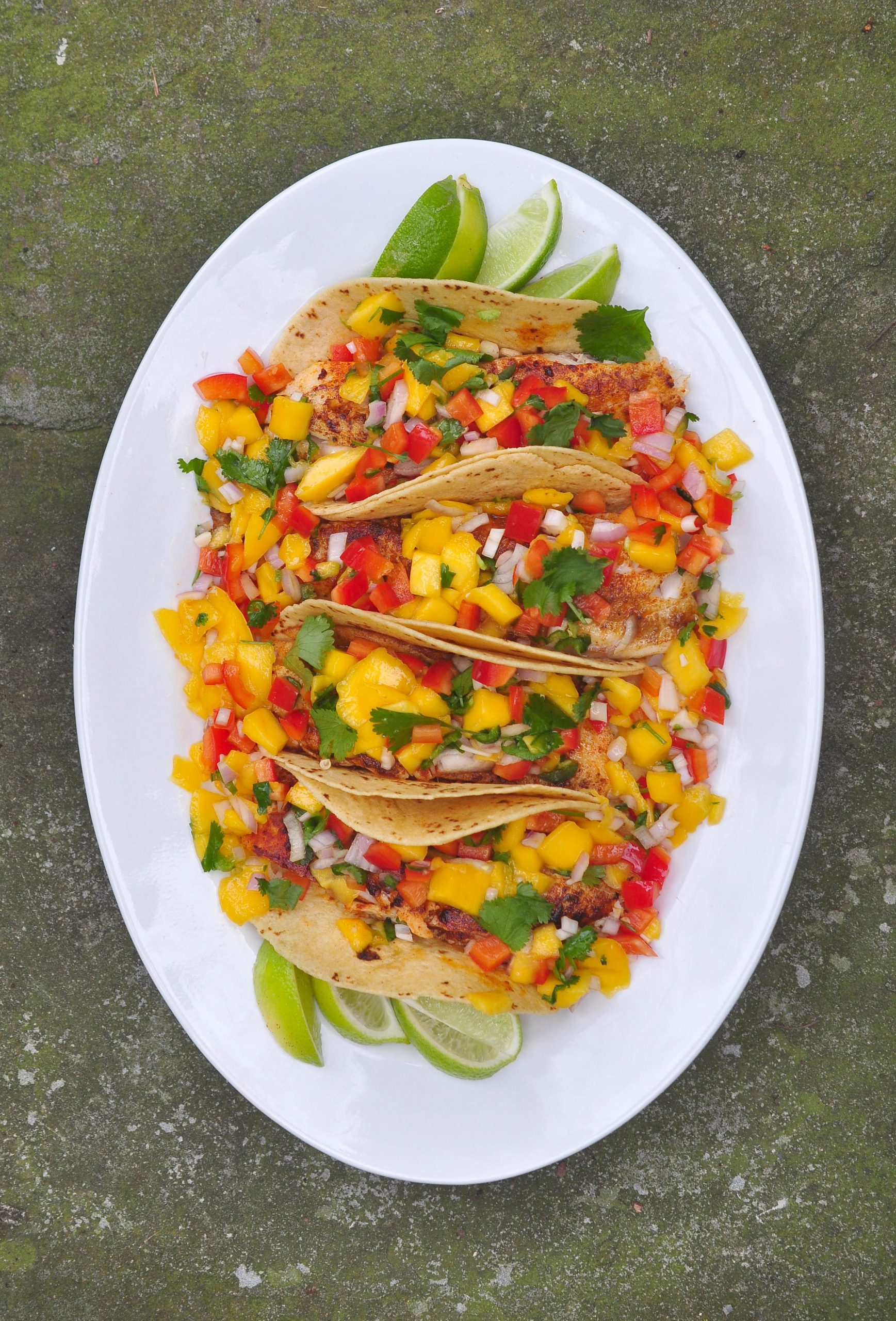 A large white oval platter with 4 tacos full of mango salsa and grilled fish. Lime wedges are nestled under the edges of the tacos. It is full of color, yellow, red and green. The plate rests on a slate background.