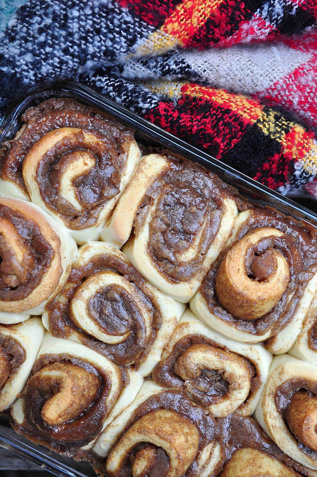 Snail-like rolls of golden brown dough bubbling over with a dark brown filling of sugary goo arranged in a baking tray with a red and green and black checked blanket nestled against the baking tray. Set at an angle.
