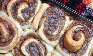 Snail-like rolls of golden brown dough bubbling over with a dark brown filling of sugary goo arranged in a baking tray with a red and green and black checked blanket nestled against the baking tray. Set at an angle.